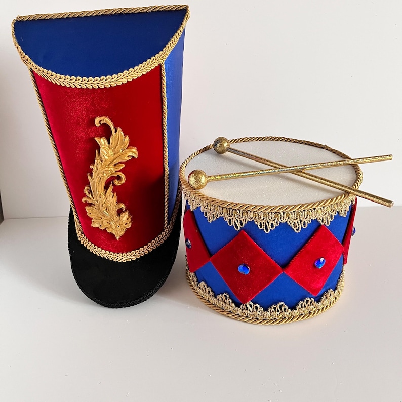 May include: A toy drum and hat set in red, blue, and gold. The drum is cylindrical with red diamond accents and blue gems. The hat is tall with a red front and gold embellishments. The set is designed to look like a marching band uniform.