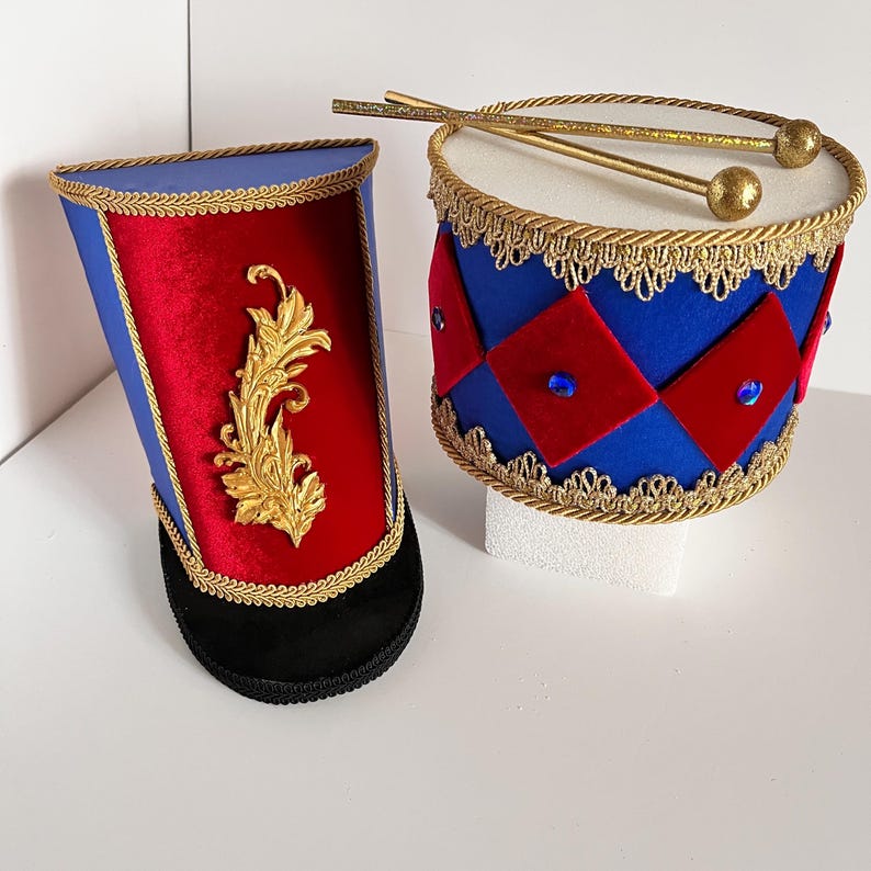 May include: A pair of decorative toy drums. One is a tall, blue and red hat-shaped drum with gold trim and a gold emblem. The other is a round drum with blue and red diamond shapes, gold trim, and drumsticks.
