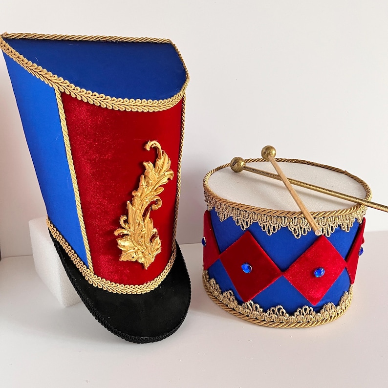 May include: A toy drum and a tall hat, likely for a child's costume. The drum is blue with red diamond accents and gold trim. The hat is blue and red with gold trim and a gold decorative element. The drumsticks are wooden.