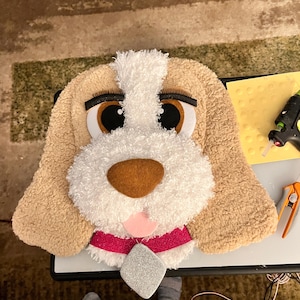 May include: A plush dog head with large brown eyes, a white muzzle, and tan ears. It has a pink collar with a silver diamond-shaped accent. The dog's face is made of soft, textured fabric.