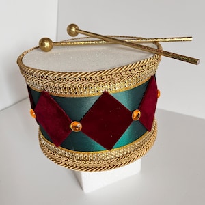 May include: A decorative drum with gold accents and drumsticks. The drum features a white top, a green body with red diamond-shaped accents, and gold trim. The drumsticks are gold with rounded ends. The drum is a festive decoration.