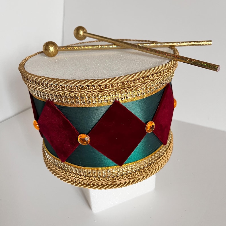May include: A decorative drum with gold accents and drumsticks. The drum features a white top, a green body with red diamond-shaped accents, and gold trim. The drumsticks are gold with rounded ends. The drum is a festive decoration.