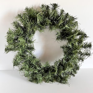 May include: A circular, artificial evergreen wreath. The wreath is composed of dense, green, needle-like foliage, creating a full and textured appearance. The wreath is designed for seasonal home decor.