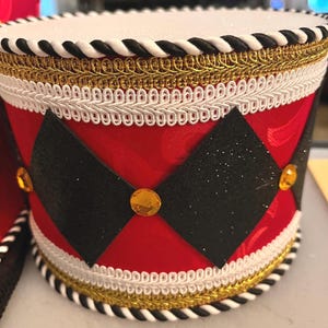 May include: A cylindrical drum-shaped decoration featuring a bright red fabric body with a subtle pattern. It is adorned with black glittery diamond shapes, gold-colored circular gems, white braided trim, and a black and white twisted rope border.