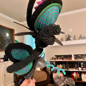May include: A whimsical, handmade decorative headpiece featuring a large, stylized flower. The flower has black, green, and purple accents, with glittery black petals and a fuzzy texture. The design includes a white and pink zig-zag pattern.
