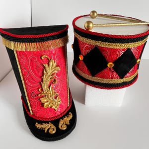 May include: Two decorative toy drums. One is a tall, black and red boot shape with gold accents and fringe. The other is a round drum with red paisley fabric, black diamond shapes, gold trim, and drumsticks.