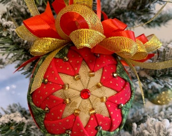 May include: A red, green, and gold Christmas ornament with a star-shaped design and a red jewel in the center. The ornament is decorated with gold ribbon and a bow.