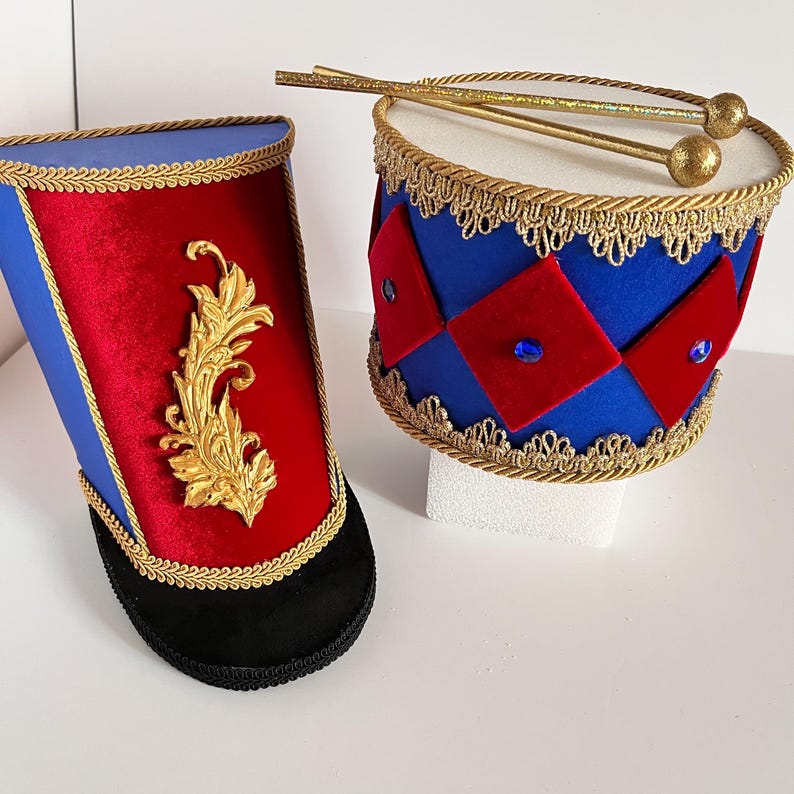 May include: A pair of decorative toy drums. One is a tall, cylindrical hat-like drum in red, blue, and gold. The other is a small drum with red and blue diamond shapes and gold trim. Drumsticks rest on top.