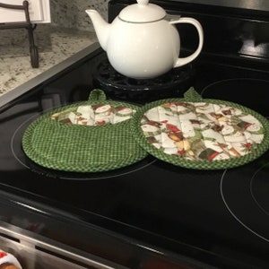 May include: Two green and white quilted pot holders with a snowman pattern. The pot holders are on a black stovetop with the words "Quick Boil" printed on the surface.