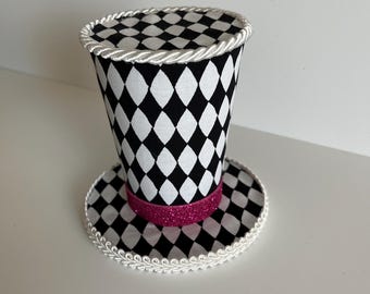 Peut inclure: Un mini chapeau haut de forme avec un motif de losanges noirs et blancs. Le chapeau est orné d'une bande rose pailletée et bordé d'une corde blanche. La base du chapeau est également à motifs de losanges.
