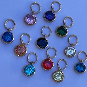 May include: Twelve gold charms featuring a variety of coloured gemstones. Each charm has a small gold loop for attaching to a bracelet or necklace. The gemstones are pink, purple, green, blue, peach, white, red, light blue, yellow, and dark blue.
