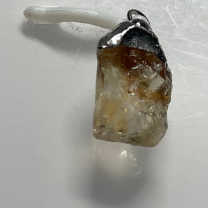 May include: A raw, light brown, crystal pendant with a silver setting. The pendant is hanging from a white string.