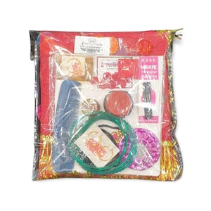 Puede incluir: Una bolsa de plástico transparente llena de varios artículos, incluyendo un peine rojo, un pequeño recipiente rojo, un brazalete verde y dorado, un brazalete rosa y dorado y una liga para el cabello negra. La bolsa también contiene una pequeña bolsa roja y dorada con el texto "ROSE MARIE The original Hairgrip Made in Germany".