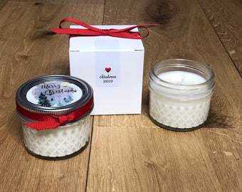 May include: A white gift box with a red ribbon and a label that says "christmas 2019" with a red heart. Next to the box is a small glass jar candle with a white label that says "Merry Christmas" with a watercolor design of a snowy forest.