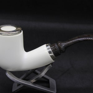 Puede incluir: Una pipa blanca con un diseño curvo y un tallo marrón oscuro. La pipa tiene detalles plateados en la cazoleta y el tallo. La pipa descansa sobre un soporte plateado sobre un fondo negro.