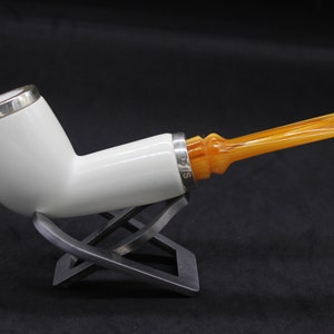 May include: A white tobacco pipe with a silver band and an amber-colored stem. The pipe is displayed on a silver stand against a dark background. The silver band has the text "S/15" engraved on it.