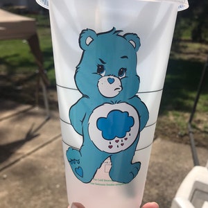 May include: A clear plastic reusable cup with a blue Care Bear character with a cloud and raindrops on it. The text on the cup reads "For Cold Beverages".