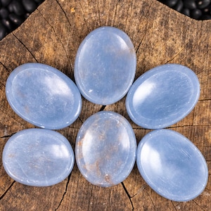 May include: Six oval-shaped, light blue stones are arranged on a weathered, brown wooden surface. The stones have a smooth, polished appearance and vary slightly in color. The wood grain is visible, adding texture to the composition.