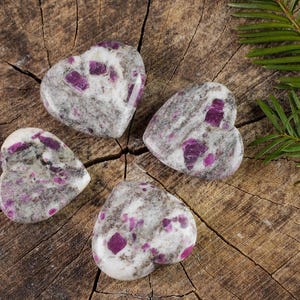 May include: Four heart-shaped polished stones with a white and gray base and pink and purple flecks. The stones are arranged in a circle on a wooden surface.