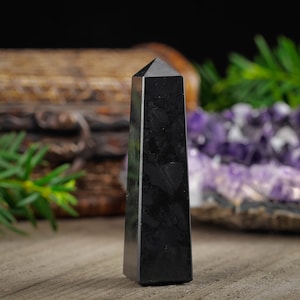 May include: A black obsidian crystal point, also known as a black obsidian obelisk, is a dark, glassy, and pointed crystal. The crystal is sitting on a wooden surface.