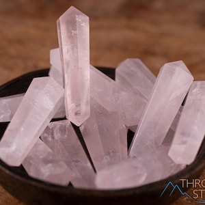May include: A collection of pale pink rose quartz points, also known as crystal wands, are arranged in a bowl. The points are smooth and polished, with a translucent appearance.