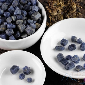 May include: Three white bowls filled with rough, blue sapphire gemstones. The bowls are arranged in a pyramid shape, with the largest bowl at the top and the smallest bowl at the bottom. The gemstones are various sizes and shapes.