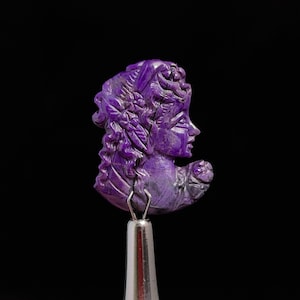 May include: A purple carved cameo of a woman's head with detailed hair and a flower in her hand.