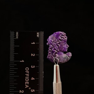 May include: A purple carved cameo of a woman's face with intricate details. The cameo is approximately 1.5 inches tall and is mounted on a silver tool.