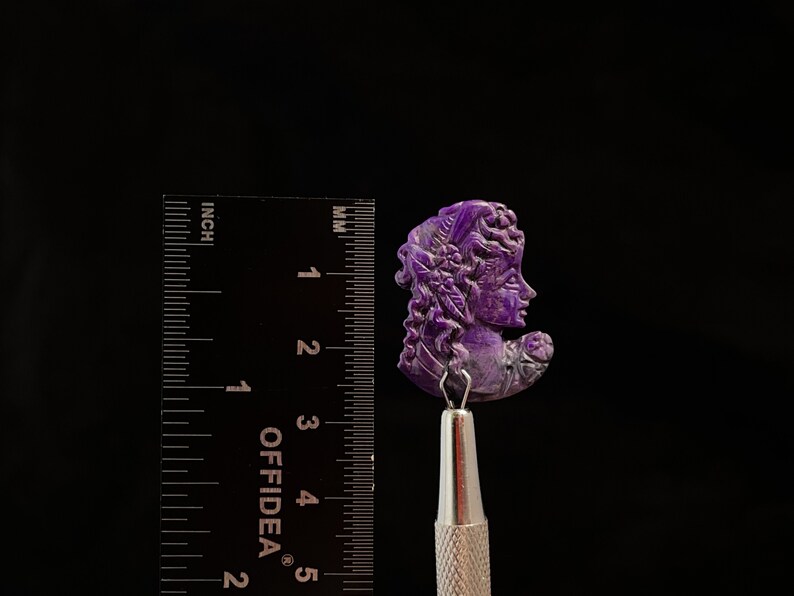 May include: A purple carved cameo of a woman's face with intricate details. The cameo is approximately 1.5 inches tall and is mounted on a silver tool.