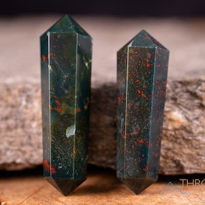 May include: Two green, red, and black gemstone points, also known as bloodstone points, are displayed on a gray rock surface. The points are polished and have a smooth, shiny finish.