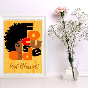 May include: A framed art print with a yellow background and the words "Focused and Blessed." The artwork features a stylized illustration of a woman with black hair, an orange earring, and red lipstick. A vase with flowers is next to the frame.