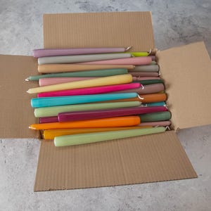 May include: A cardboard box filled with a variety of taper candles in assorted colors, including purple, pink, yellow, green, blue, orange, and brown.