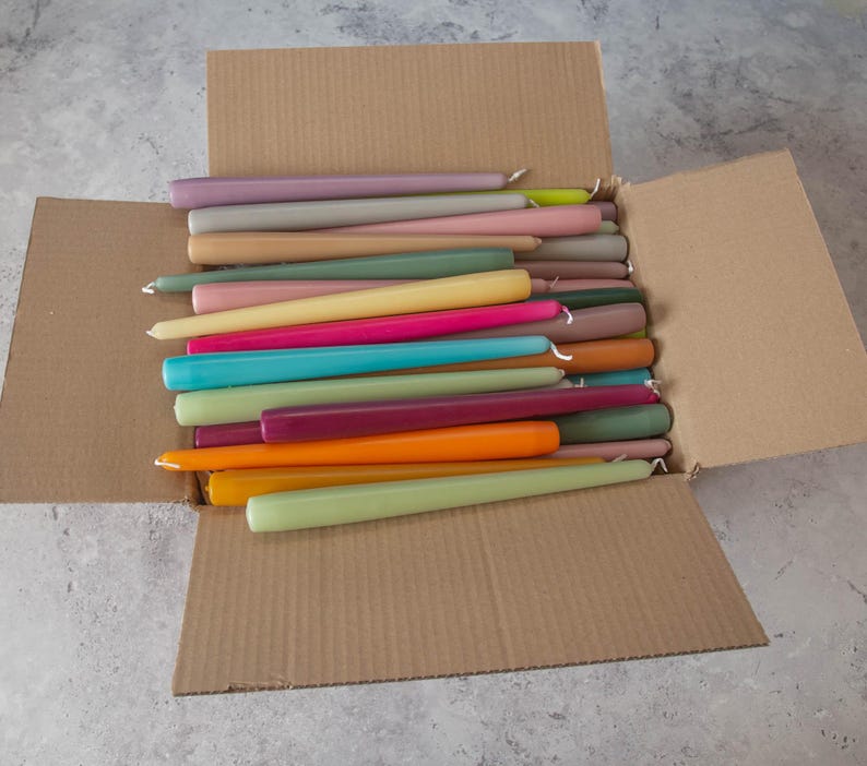 May include: A cardboard box filled with a variety of taper candles in assorted colors, including purple, pink, yellow, green, blue, orange, and brown.