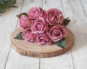 May include: A bouquet of six pink silk roses arranged on a wooden slice. The roses are clustered together and have a soft, velvety texture.