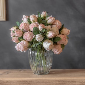 May include: A clear glass vase with a ribbed design, filled with a bouquet of pink roses and green foliage. The roses are in different stages of bloom. The vase is placed on a wooden surface, set against a dark grey backdrop.