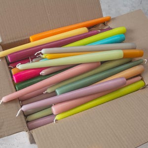 May include: A box filled with a variety of colorful taper candles. The candles are arranged in a random pattern and are all the same size. The candles are in shades of pink, green, yellow, orange, and purple.