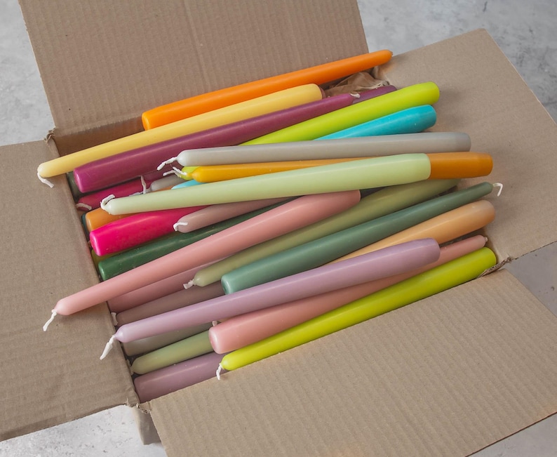 May include: A box filled with a variety of colorful taper candles. The candles are arranged in a random pattern and are all the same size. The candles are in shades of pink, green, yellow, orange, and purple.
