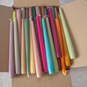 May include: A box filled with a variety of taper candles in different colors, including purple, gray, beige, green, yellow, pink, red, orange, blue, and brown.