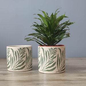 May include: Two cream-coloured ceramic plant pots with a green leaf pattern. One pot is empty, and the other contains a green plant. The pots are cylindrical and sit on a wooden surface.