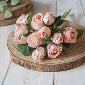 May include: A bouquet of blush pink silk roses arranged on a wooden slice. The roses are clustered together and have a realistic appearance.