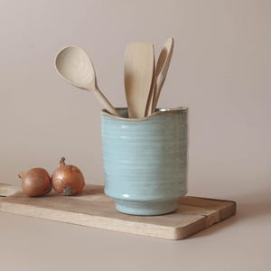 May include: A pale blue ceramic utensil holder containing wooden spoons and spatulas. Two onions rest on a wooden chopping board. The holder has a textured finish and a wavy edge. The spoons and spatulas are made of light-coloured wood.