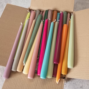 May include: A box filled with a variety of taper candles in different colors, including purple, green, blue, pink, orange, and yellow.