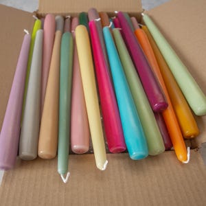 May include: A collection of long, thin taper candles in a variety of colors, including purple, gray, beige, green, pink, yellow, blue, and orange. The candles are arranged in a box.