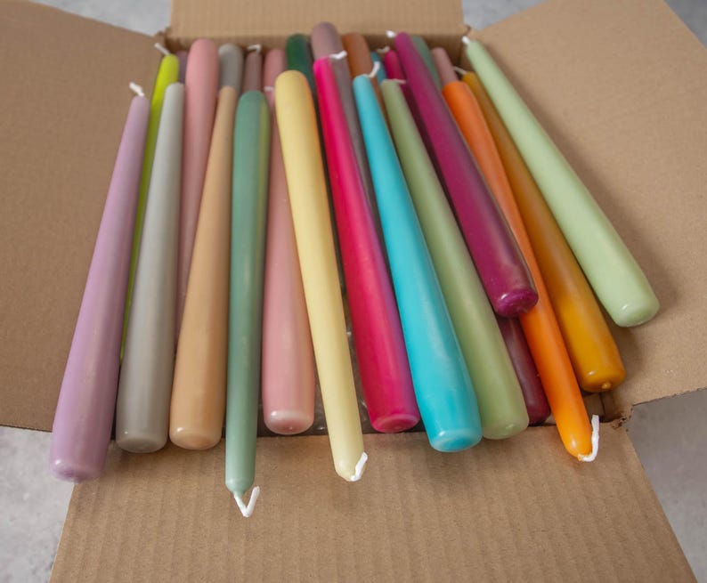 May include: A collection of long, thin taper candles in a variety of colors, including purple, gray, beige, green, pink, yellow, blue, and orange. The candles are arranged in a box.