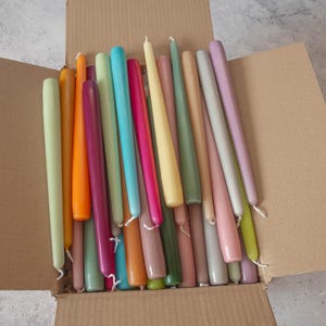 May include: A box filled with a variety of long, thin taper candles in a range of pastel colors, including light green, pink, yellow, orange, and blue.