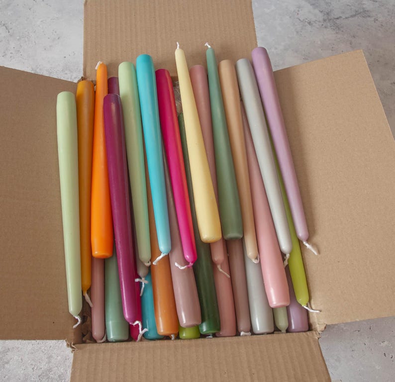 May include: A box filled with a variety of long, thin taper candles in a range of pastel colors, including light green, pink, yellow, orange, and blue.