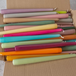 May include: A box filled with a variety of taper candles in assorted colors, including purple, pink, beige, yellow, green, blue, orange, and red.