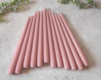 May include: A set of 12 long, thin, dusty rose taper candles. The candles are arranged in a fan shape on a gray surface.