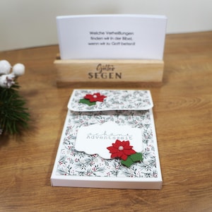 Può includere: Una confezione regalo decorativa a tema natalizio con un motivo floreale e un accento di stella di Natale rossa. La scatola ha il testo "Schöne Adventszeit" sul davanti. Un supporto in legno con il testo "Gottes Segen" contiene una carta con testo.