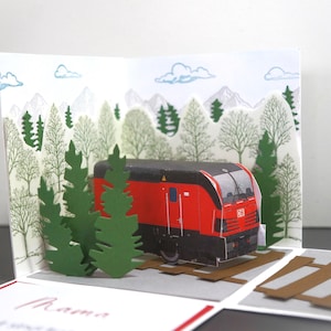Pode incluir: Um cartão pop-up com um comboio vermelho e preto sobre carris, com um fundo de árvores e montanhas. Árvores de papel verde em primeiro plano. O cartão tem um tema florestal.